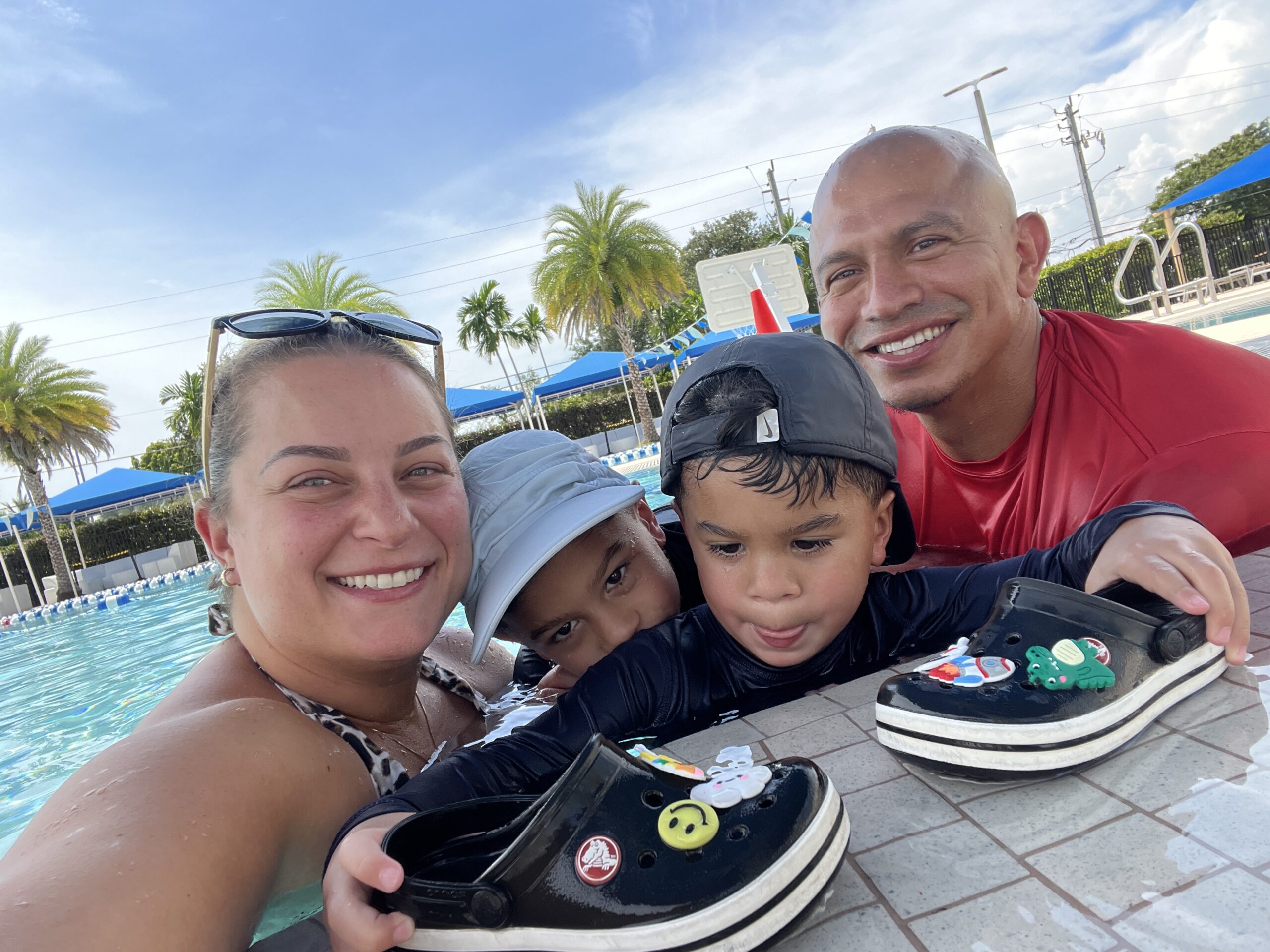 A family of four at the pool: mom and dad sitting poolside, smiling and watching over their two young sons who are playing in the water. The scene captures a joyful, relaxed family moment in the sun.